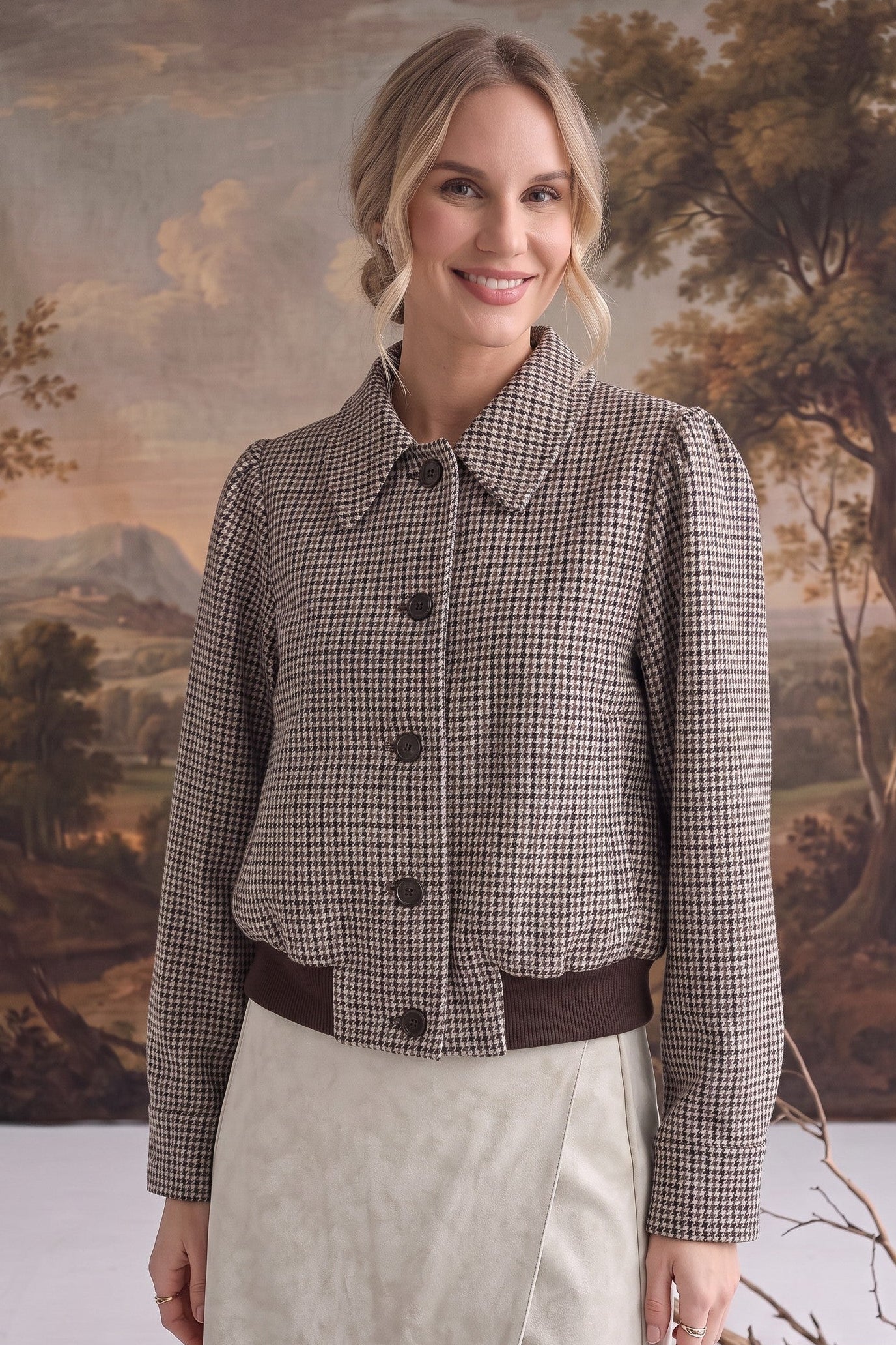 A woman modeling a cropped, long-sleeve button-front jacket with a brown and beige houndstooth pattern, paired with a cream skirt, set against a rustic landscape backdrop.