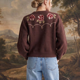 A woman stands with her back to the camera wearing a brown knit sweater with embroidered pink and red roses on the back, paired with light blue wide-leg jeans. She is posed in front of a large landscape tapestry with decorative branches on the floor.