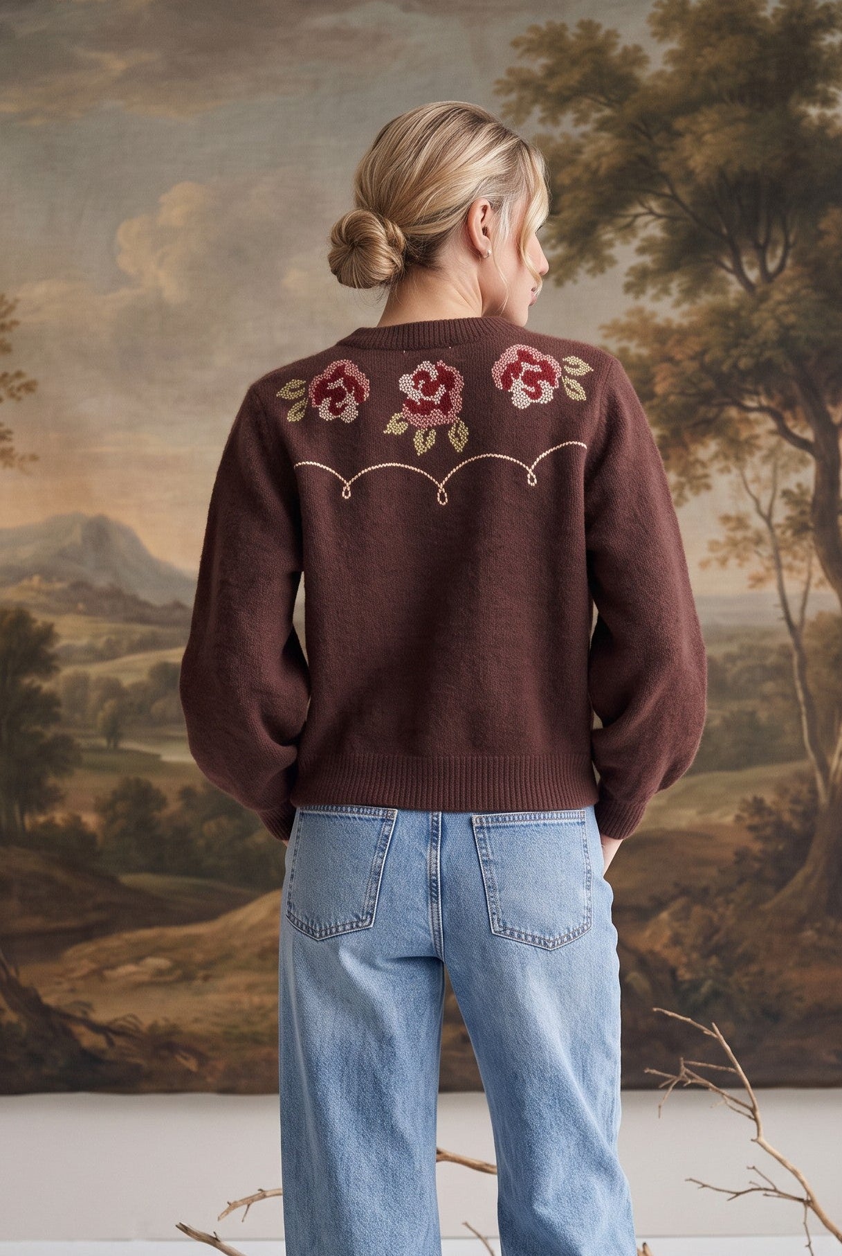 A woman stands with her back to the camera wearing a brown knit sweater with embroidered pink and red roses on the back, paired with light blue wide-leg jeans. She is posed in front of a large landscape tapestry with decorative branches on the floor.