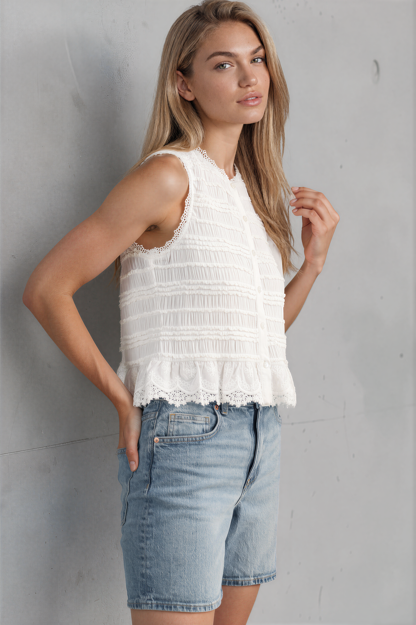 Woman wearing a white sleeveless top and denim shorts against a gray wall.