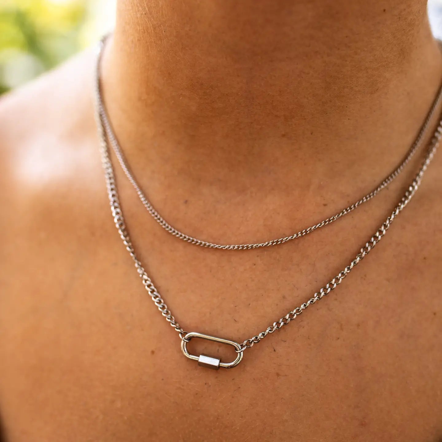 A close-up image of a silver necklace with a small, rectangular pendant, worn on a person's neck.