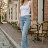 Woman in white tank top and blue jeans standing on a street with brick pavement and buildings in the background.