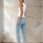 Woman wearing a white tank top and light blue jeans against a textured wall.