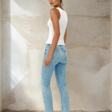 Woman wearing a white tank top and light blue jeans against a textured wall.