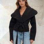 Woman wearing a black coat with a belt against a textured wall.
