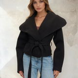 Woman wearing a black coat with a belt against a textured wall.