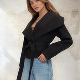 Woman wearing a black coat and blue jeans against a textured wall.