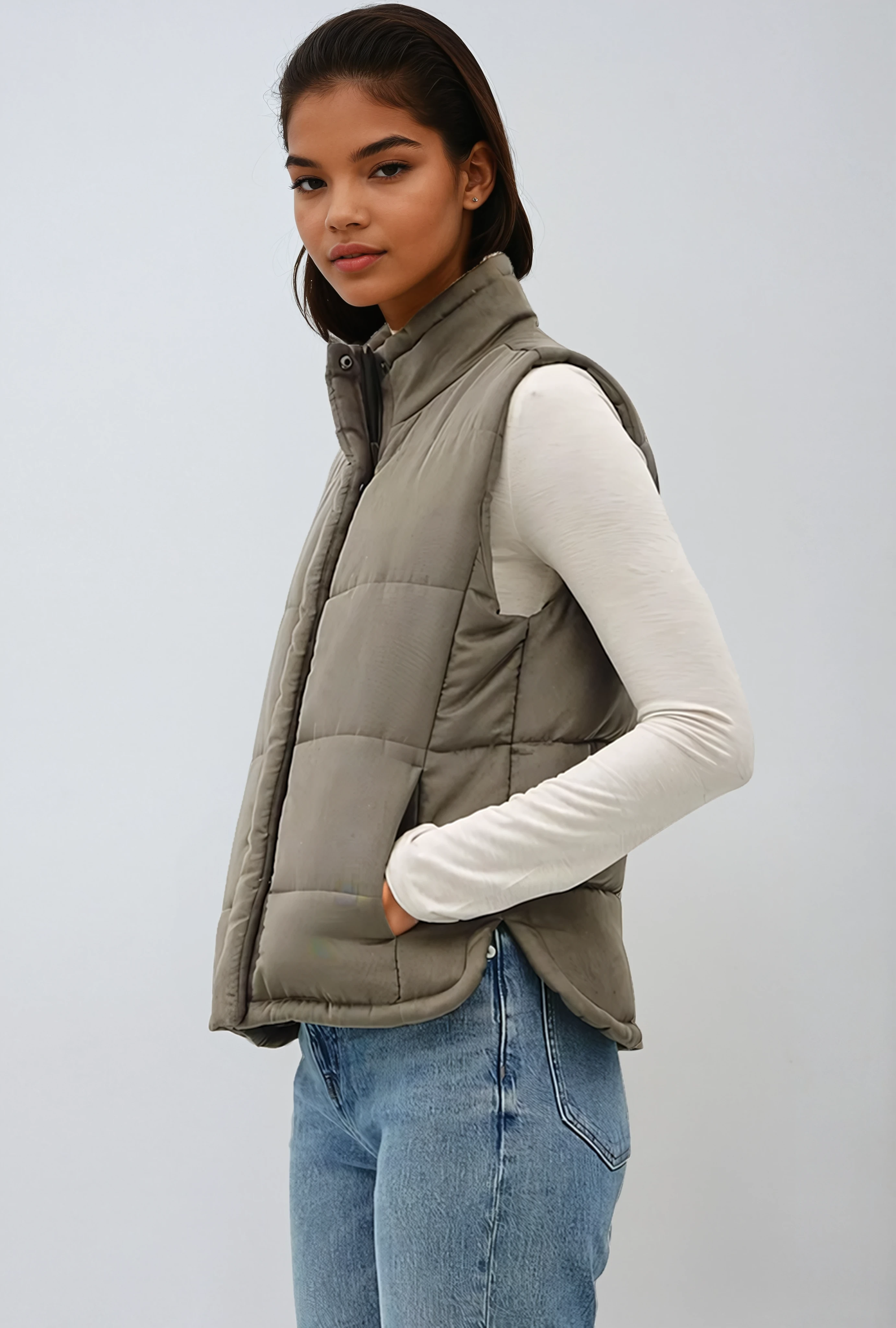 Person wearing a beige puffer vest over a white shirt with blue jeans on a light gray background