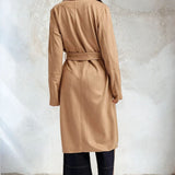 Woman wearing a beige trench coat against a textured white wall.