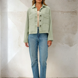Woman wearing a light green jacket and blue jeans against a textured wall.