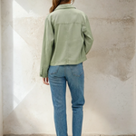 Person wearing a green jacket and blue jeans standing against a textured white wall.