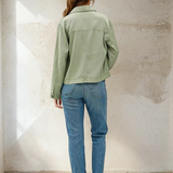 Person wearing a green jacket and blue jeans standing against a textured white wall.