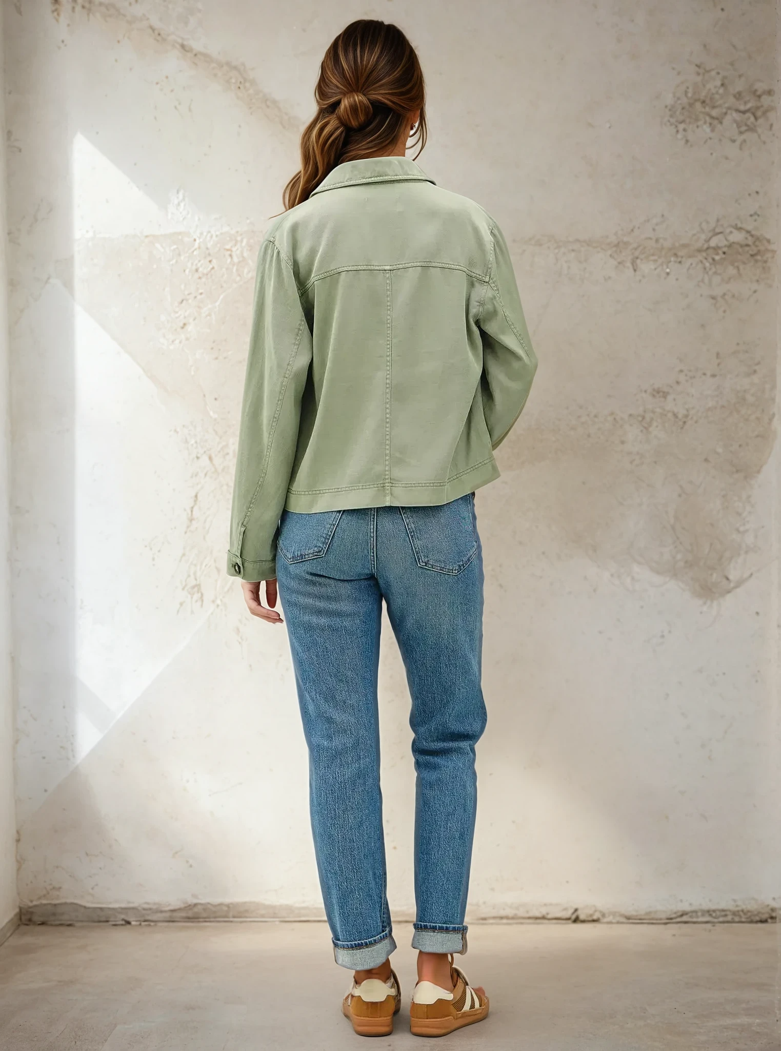 Person wearing a green jacket and blue jeans standing against a textured white wall.