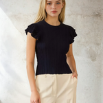 Woman wearing a black ruffled top and beige pants against a textured wall.