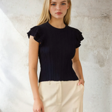 Woman wearing a black ruffled top and beige pants against a textured wall.