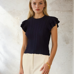 Woman wearing a black ruffled top and white pants against a light background
