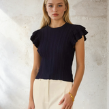 Woman wearing a black ruffled top and white pants against a light background
