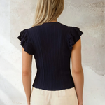 Woman wearing a black ruffled top and beige pants against a textured wall.