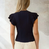 Woman wearing a black ruffled top and beige pants against a textured wall.