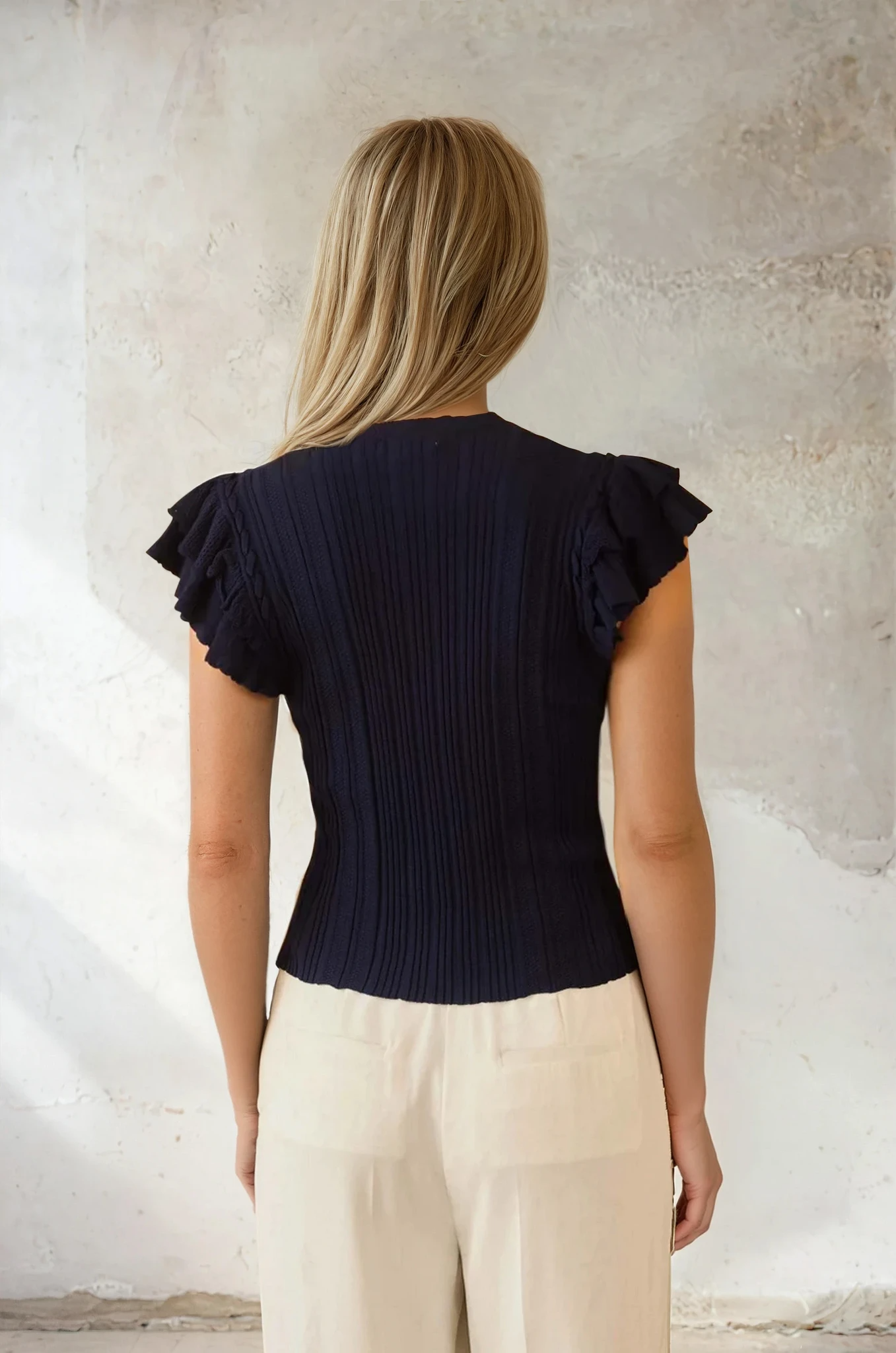 Woman wearing a black ruffled top and beige pants against a textured wall.