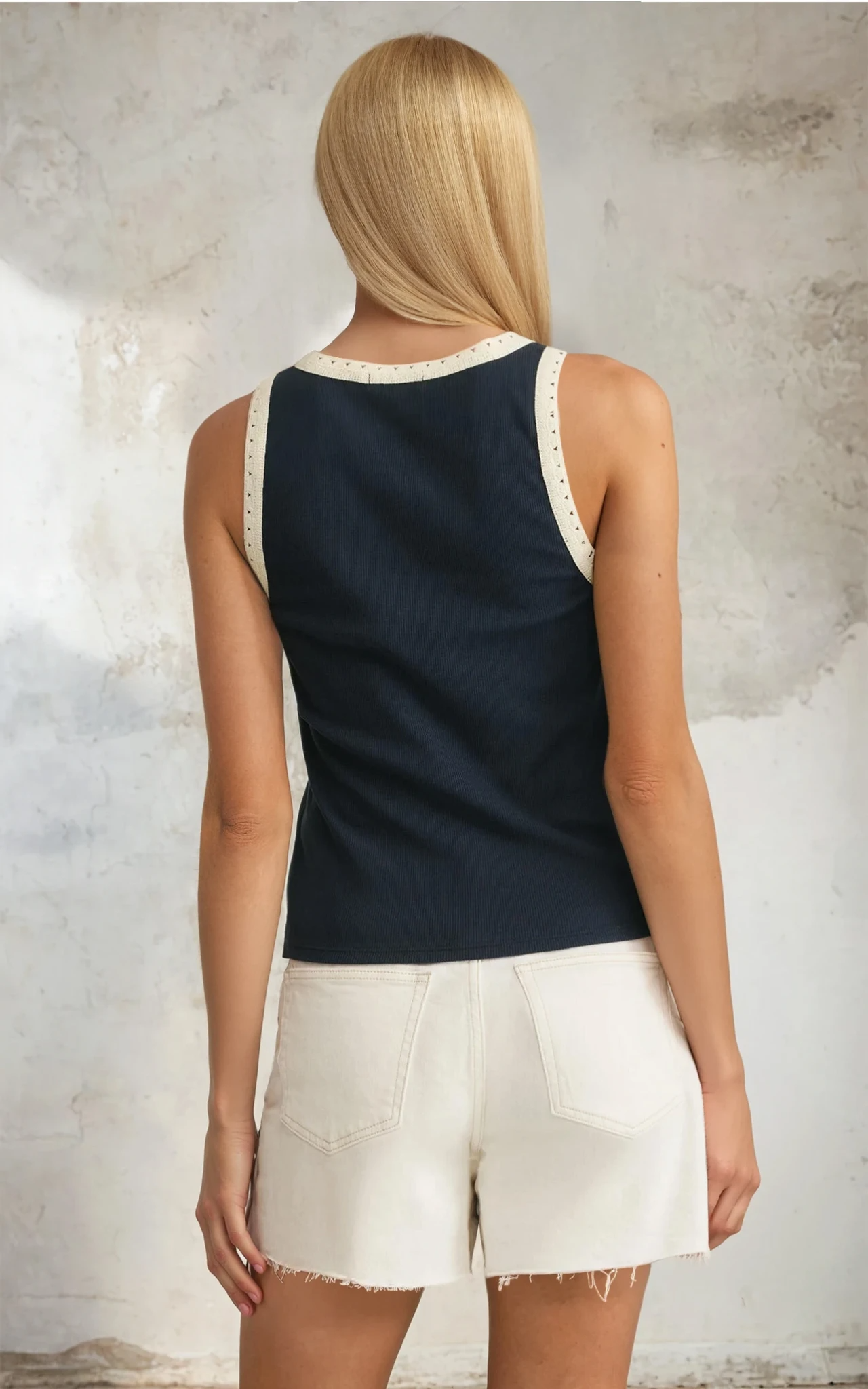 Woman wearing a navy tank top with white trim and white shorts against a textured wall.