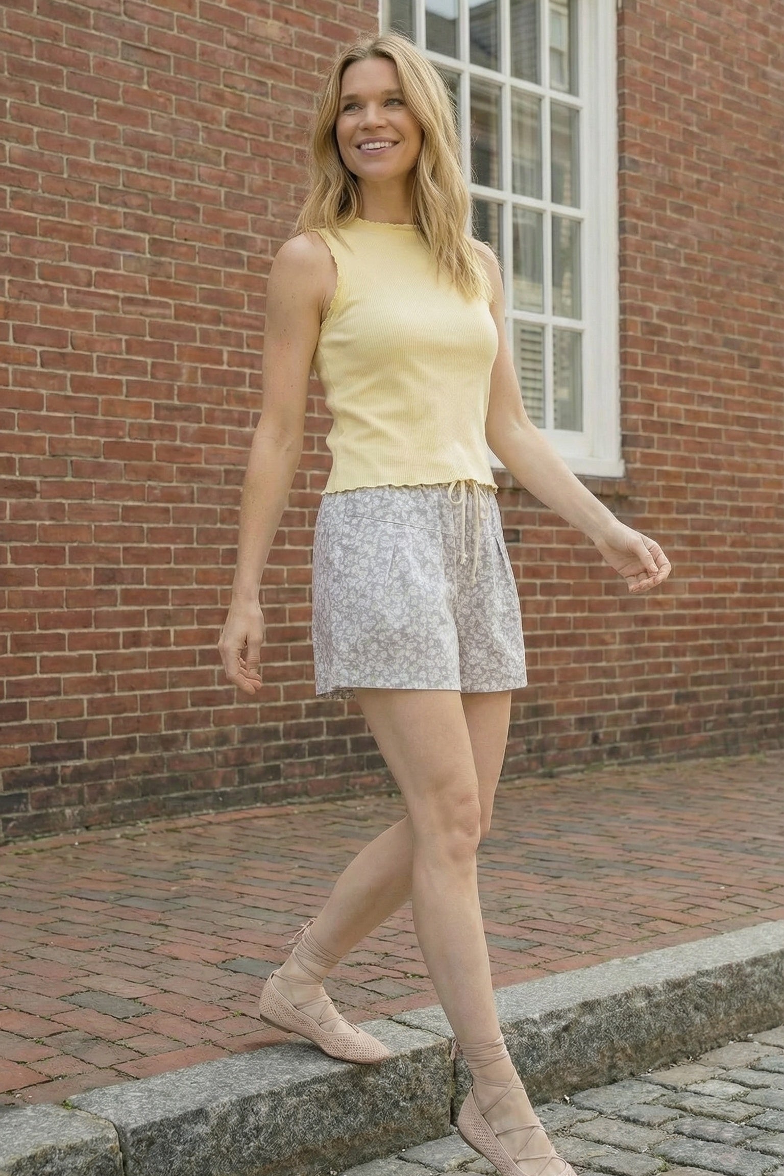 Woman in yellow top and patterned shorts walking on a sidewalk with a brick building in the background