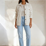 Woman wearing a beige jacket, white sweater, light blue jeans, and brown boots against a white background