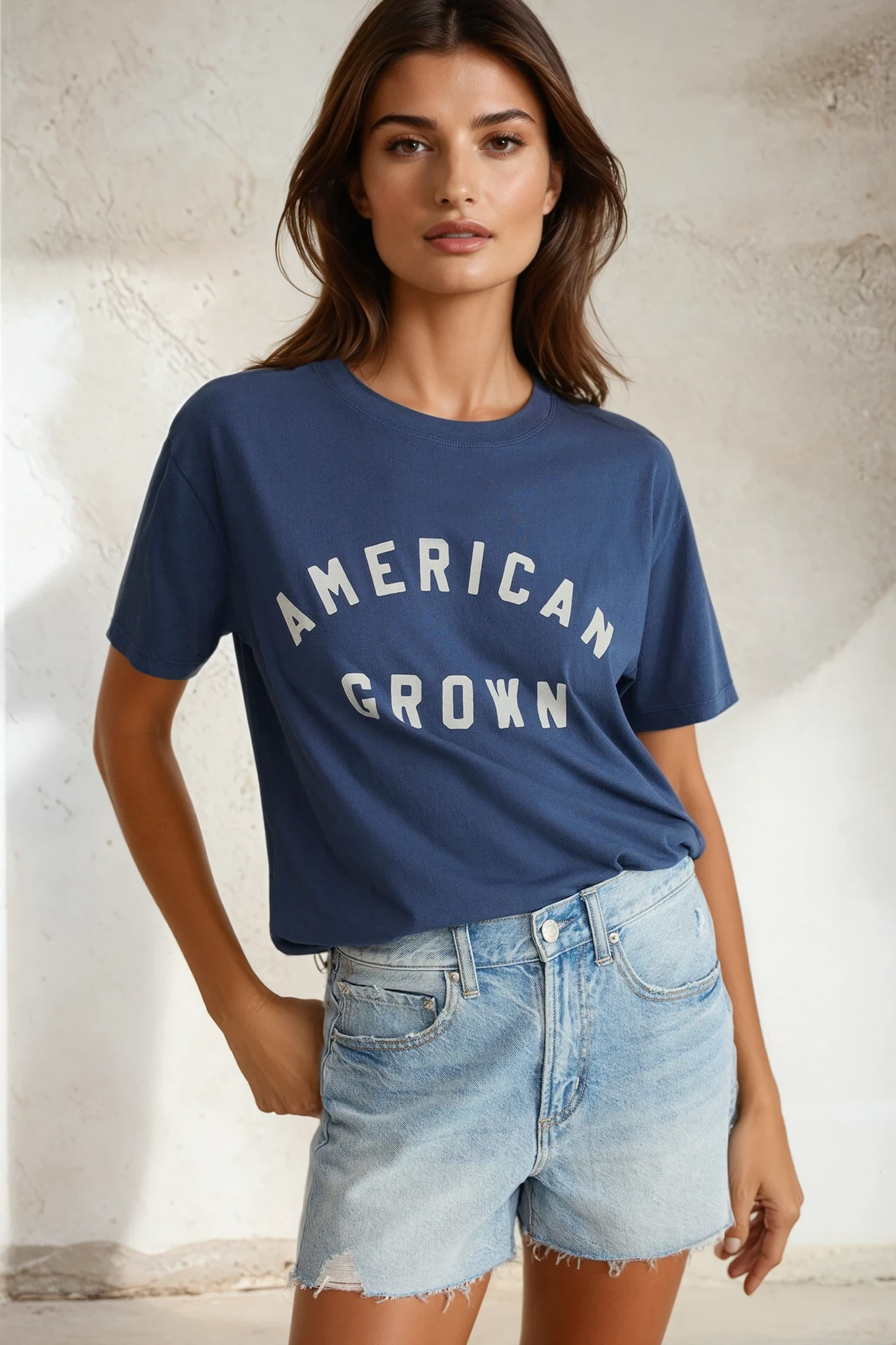 Woman wearing a blue 'American Grown' t-shirt and denim shorts against a neutral background