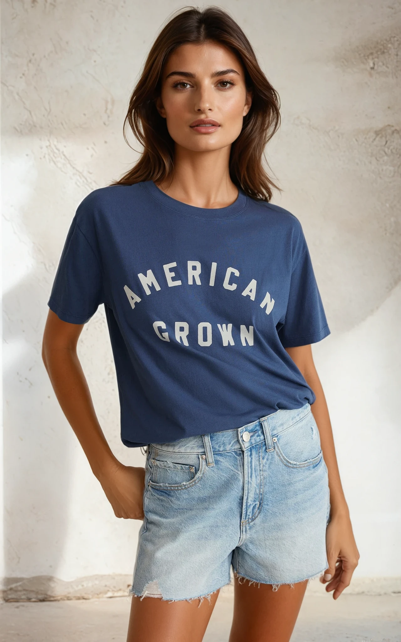 Woman wearing a blue 'American Grown' t-shirt and denim shorts against a neutral background