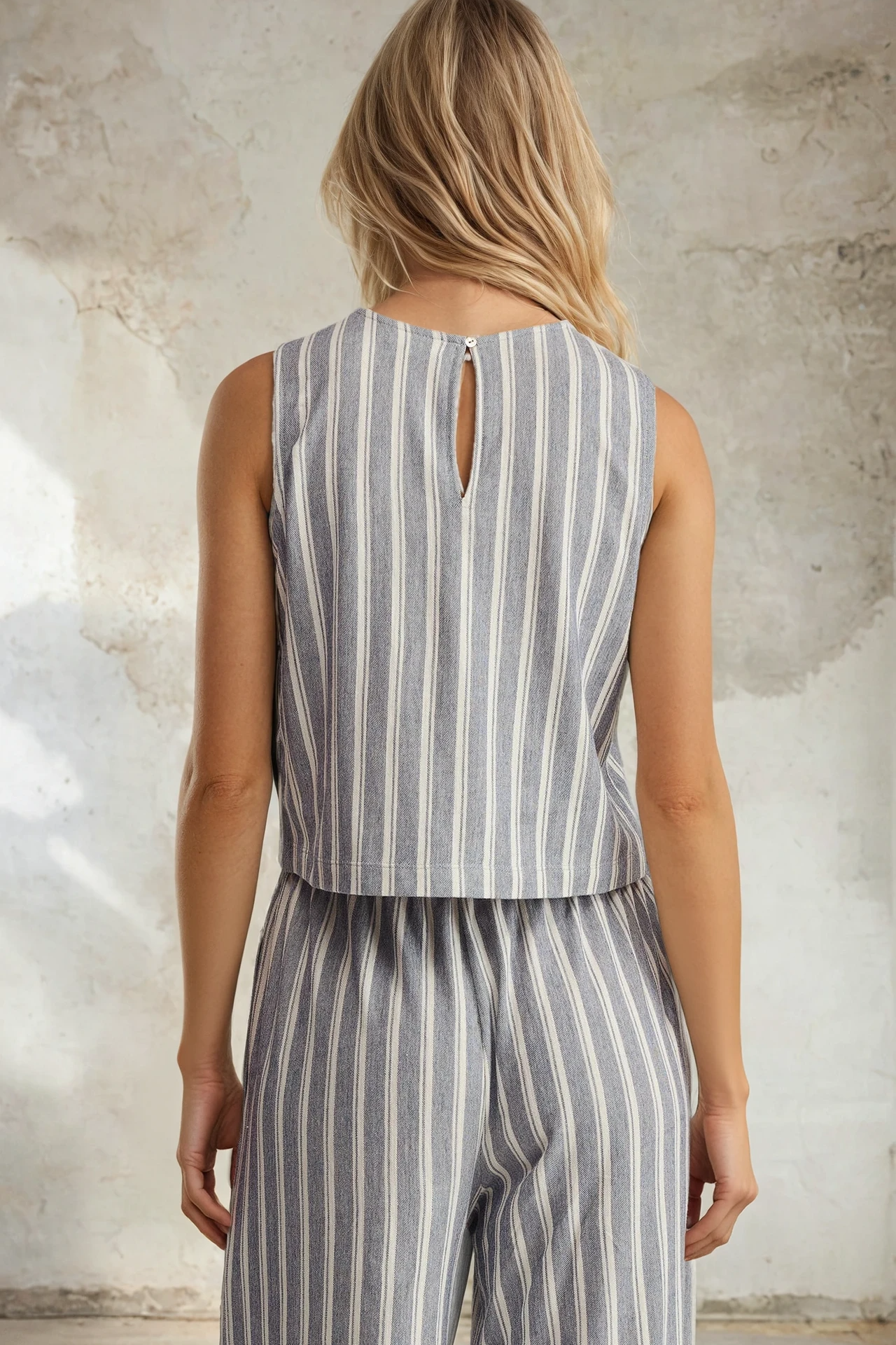 Person wearing a sleeveless striped top and pants against a textured wall.