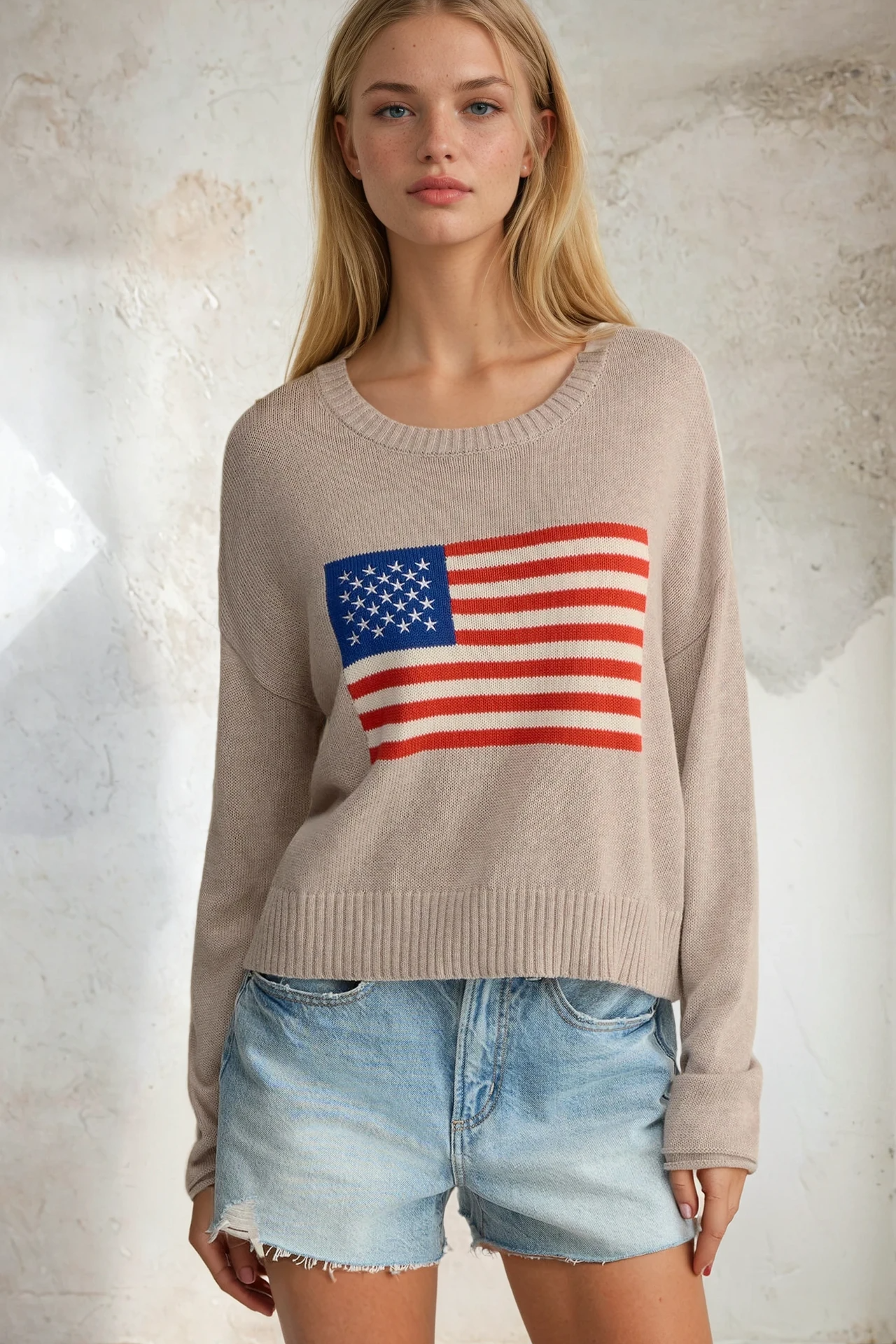 Woman wearing a beige sweater with an American flag design against a textured wall.