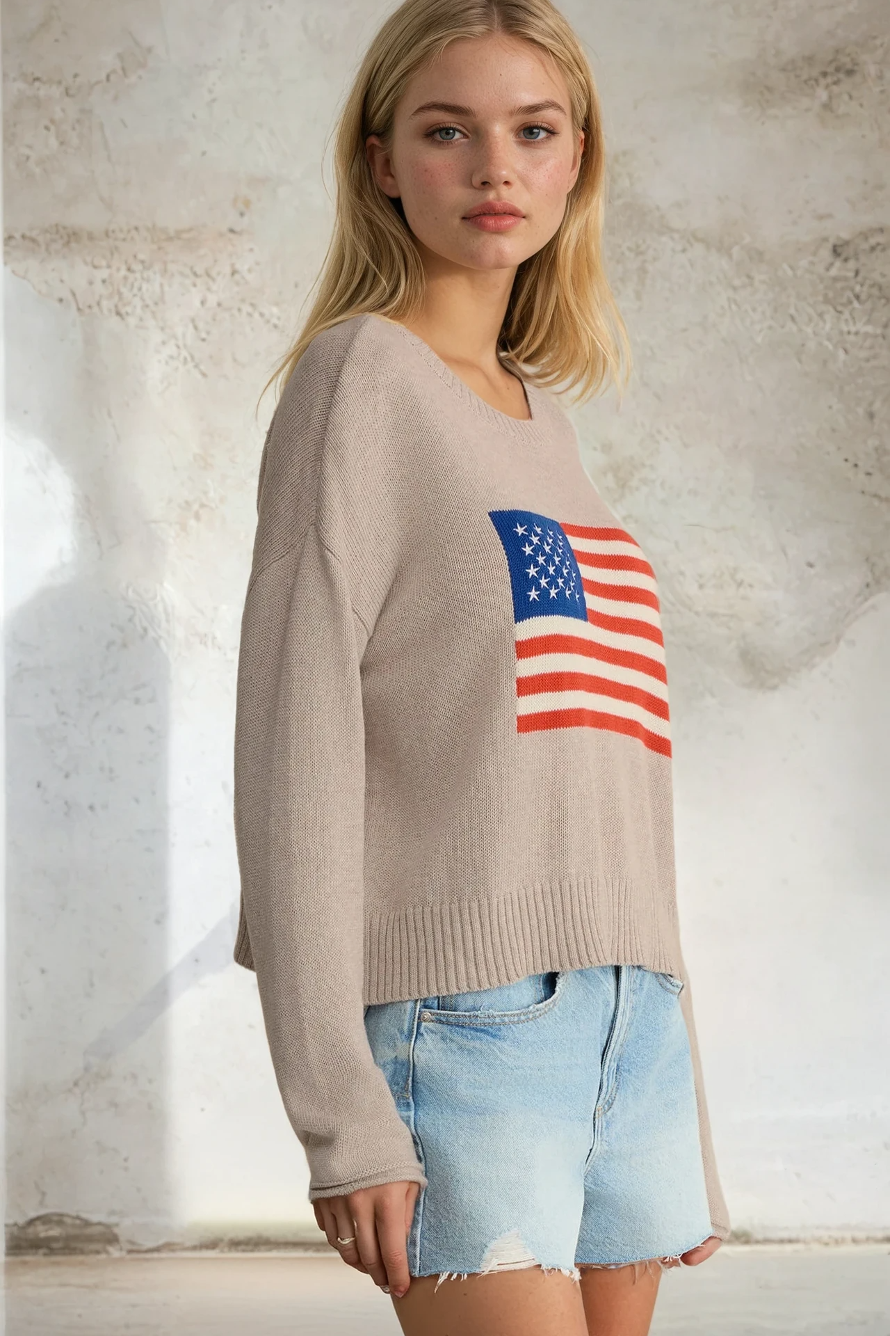 Woman wearing a beige sweater with an American flag design against a textured wall.