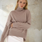 Woman wearing a beige turtleneck sweater against a textured wall.