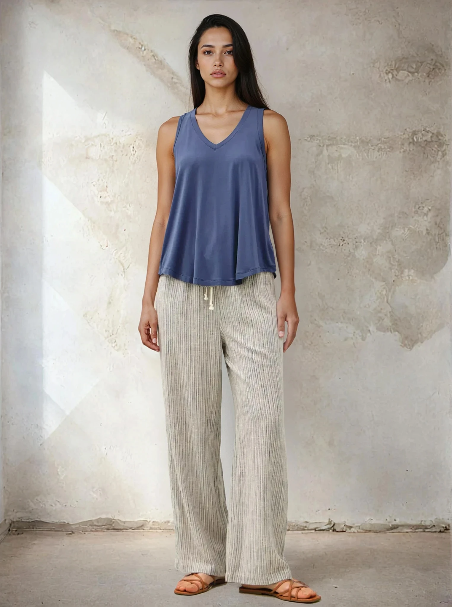 Woman wearing a blue sleeveless top and light-colored pants against a textured wall.