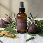 An amber glass spray bottle labeled 'aracet' and 'BROWN ORGANIC & NATURAL' stands on a white linen cloth, surrounded by figs, olive branches, and a small pile of brown powder against a softly lit neutral wall.