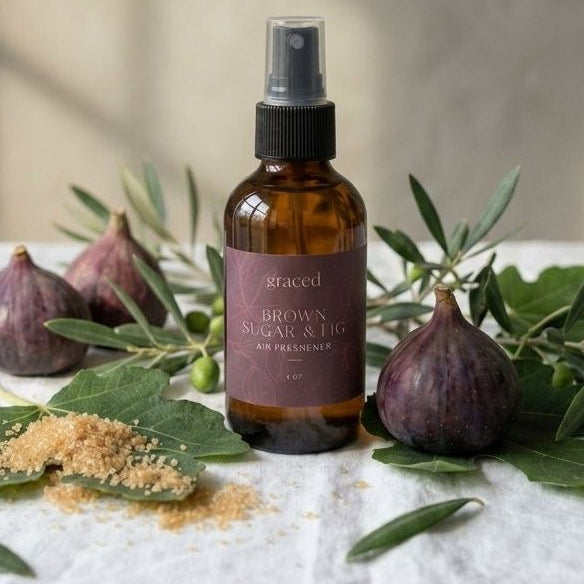 An amber glass spray bottle labeled 'aracet' and 'BROWN ORGANIC & NATURAL' stands on a white linen cloth, surrounded by figs, olive branches, and a small pile of brown powder against a softly lit neutral wall.