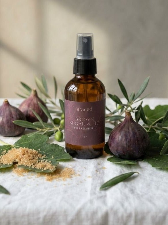An amber glass spray bottle labeled 'aracet' and 'BROWN ORGANIC & NATURAL' stands on a white linen cloth, surrounded by figs, olive branches, and a small pile of brown powder against a softly lit neutral wall.