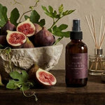 A dark amber spray bottle with a black nozzle and a purple label sits on a rustic wooden table beside a bowl of fresh figs, green leaves, a reed diffuser, and a bar of soap against a textured beige wall.