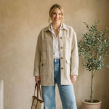 Woman wearing a beige jacket and blue jeans standing in a room with a plant and white sofa.