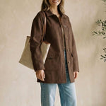 Woman in a brown coat and blue jeans standing indoors with a plant in the background