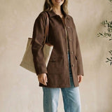 Woman in a brown coat and blue jeans standing indoors with a plant in the background