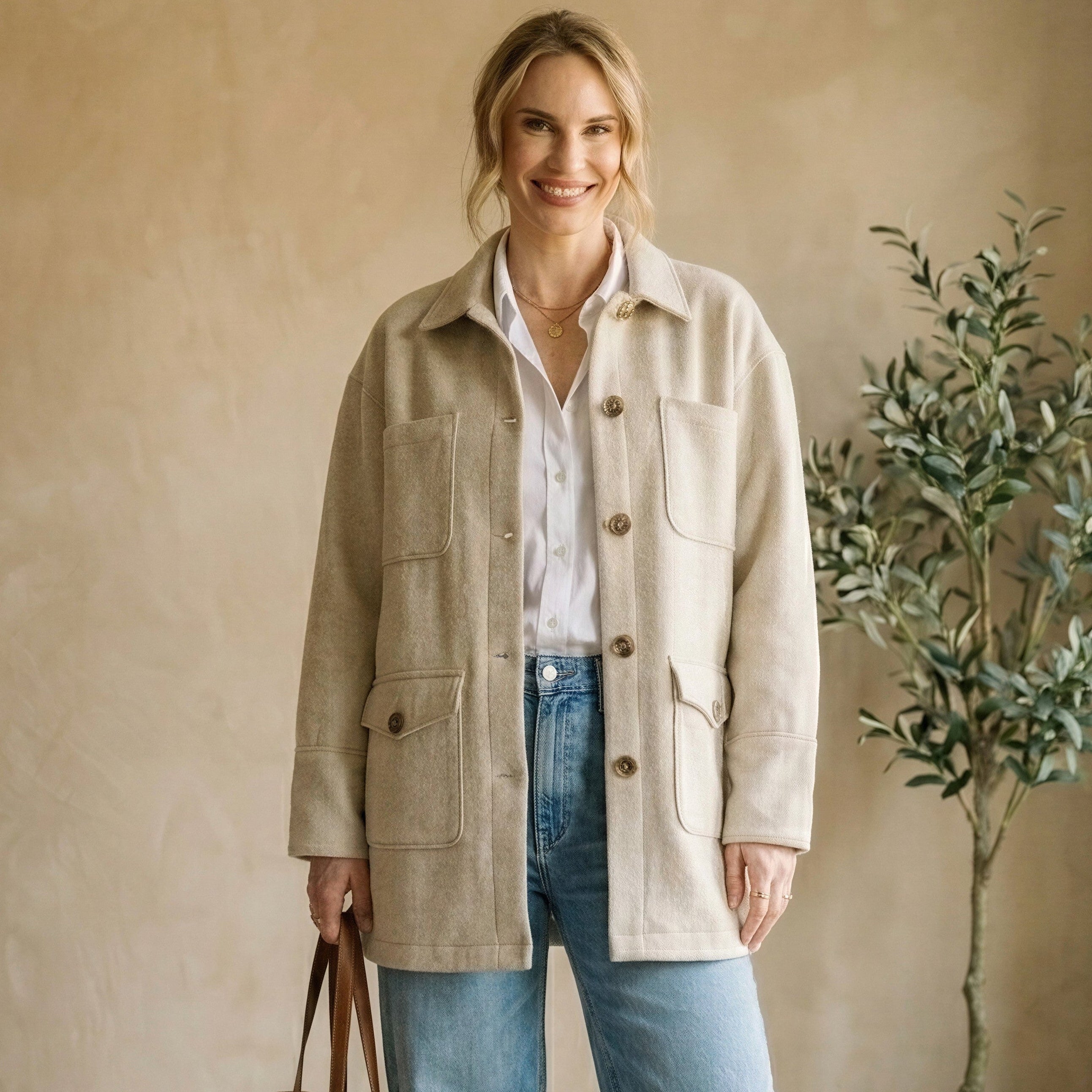 Woman wearing a beige jacket and blue jeans standing in a room with a plant and white sofa.