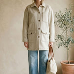 Woman wearing a beige coat and blue jeans standing in a room with a plant and bag.