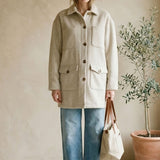 Woman wearing a beige coat and blue jeans standing in a room with a plant and bag.