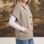 Woman wearing a graced taupe button-down hooded vest over a white long-sleeve shirt and blue jeans