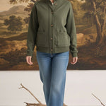 A woman wearing a graced olive green mock neck button-down cardigan with front pockets, paired with blue jeans.