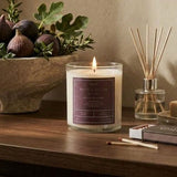 A lit candle in a clear glass jar sits on a wooden table beside a reed diffuser, soap bar, matches, and a stone bowl filled with figs and greenery against a warm beige wall.
