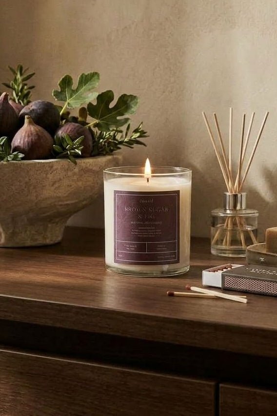 A lit candle in a clear glass jar sits on a wooden table beside a reed diffuser, soap bar, matches, and a stone bowl filled with figs and greenery against a warm beige wall.
