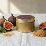 A gold metal tin with a purple lid sits on a white linen surface, styled with whole and halved figs, mint leaves, and small piles of the product's brown powder. The background is a textured beige wall with soft window light shadows.