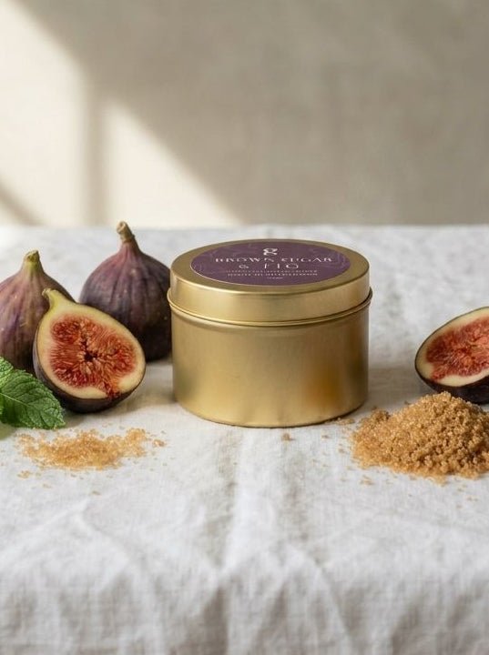 A gold metal tin with a purple lid sits on a white linen surface, styled with whole and halved figs, mint leaves, and small piles of the product's brown powder. The background is a textured beige wall with soft window light shadows.